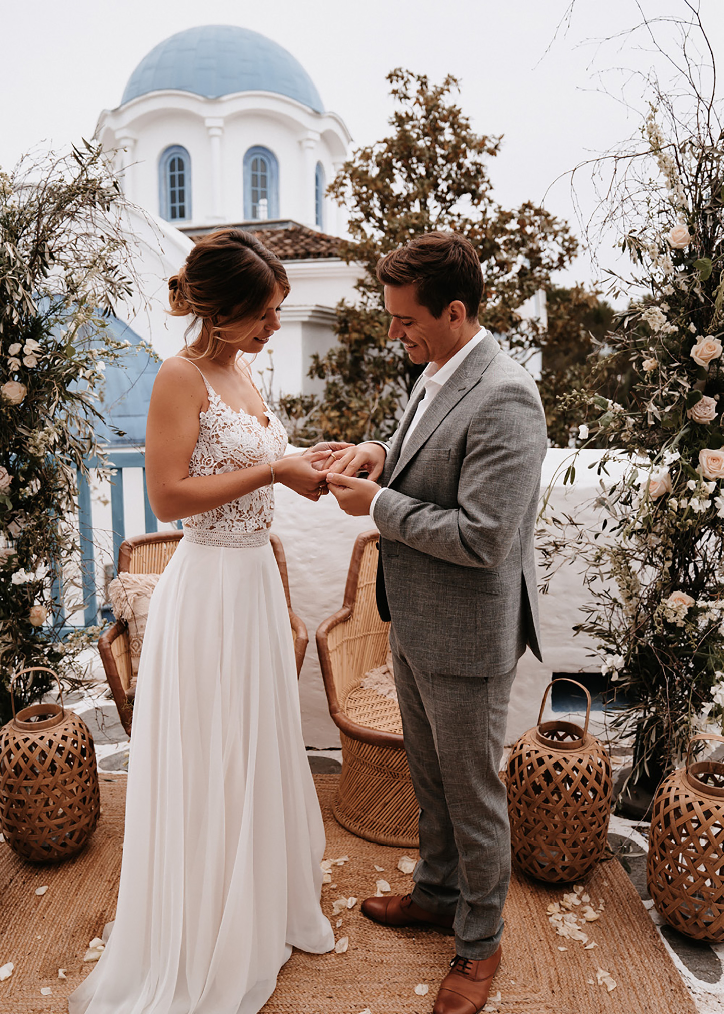 Vor dem Anblick einer griechischen Kapelle mit blauem Dach und geschmückter Atmosphäre steckt die Braut dem Bräutigam den Ehering an den Finger. 