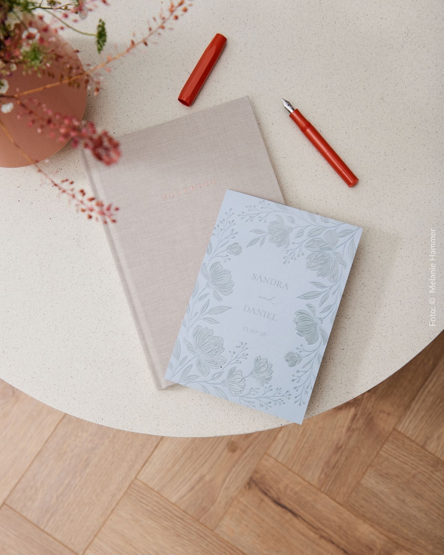 Hochzeitseinladung mit Notizbuch und Füller mit Stoffeinband auf rundem Tisch mit Blumendeko