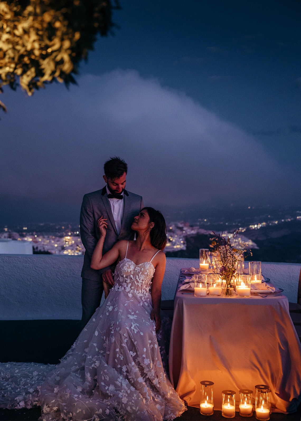 Das Brautpaar sitzt in der Dämmerung vor dem Hochzeitstisch und hinter ihnen sieht man die Lichter der Orte