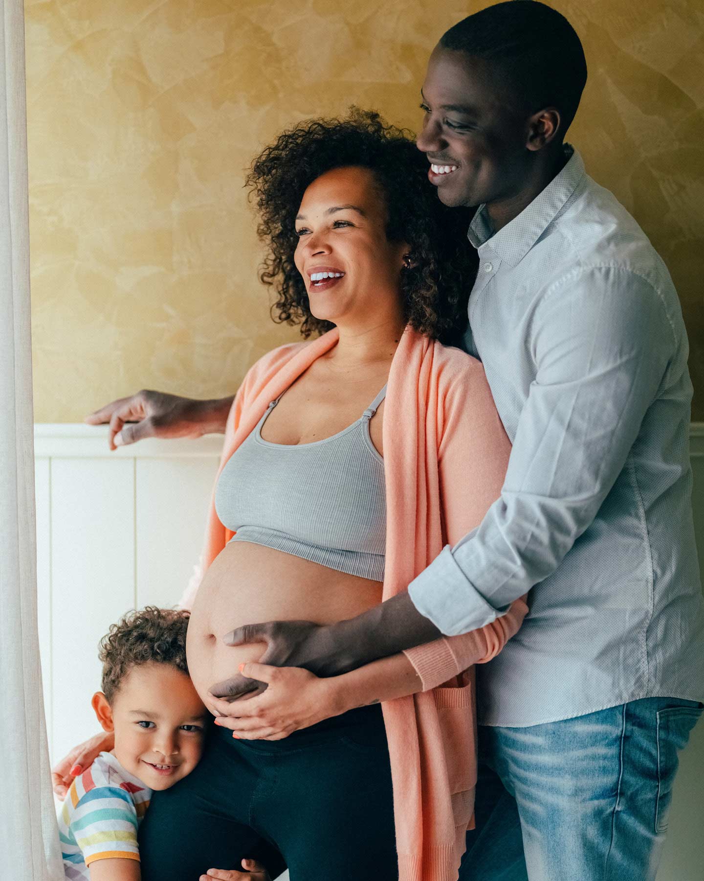 Papa hält Bauch der hochschwangeren Mama, während das Kind sich an das Bein der Mama kuschelt