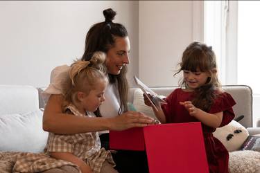 Frau auf der Couch mit 2 kleinen Mädchen öffnet ihr Weihnachtsgeschenk
