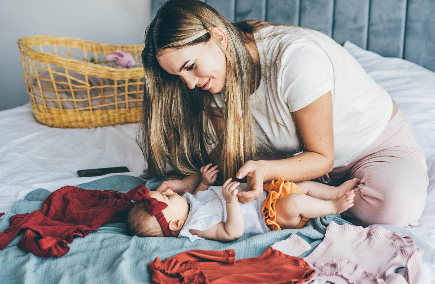 Mama auf Bett probiert ihrem Baby verschiedene Second Hand Sachen an, die Baby Kosten sparen.