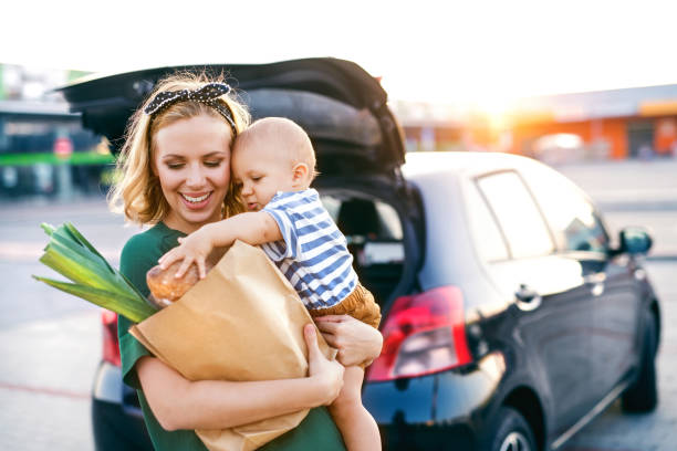 Mama mit Baby und Einkaufstüte auf dem Arm vor Auto mit geöffneter Heckklappe