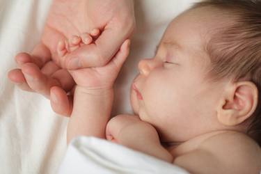 Nahaufnahme von Baby auf weißer Decke, seine Hand hält den Daumen des Vaters (nicht imBild)