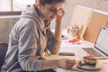 Young women celebrate birthday in living room and eating birthday cake. Women sitting at desk and work on some task on laptop.