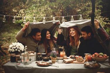   Freunde sitzen am gedeckten Tisch im Garten. Über ihren Köpfen halten sie eine Decke, um sich vor dem Regen zu schützen.