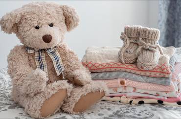 Teddybär neben einem Stapel niedlicher Babykleidung auf einem Bett