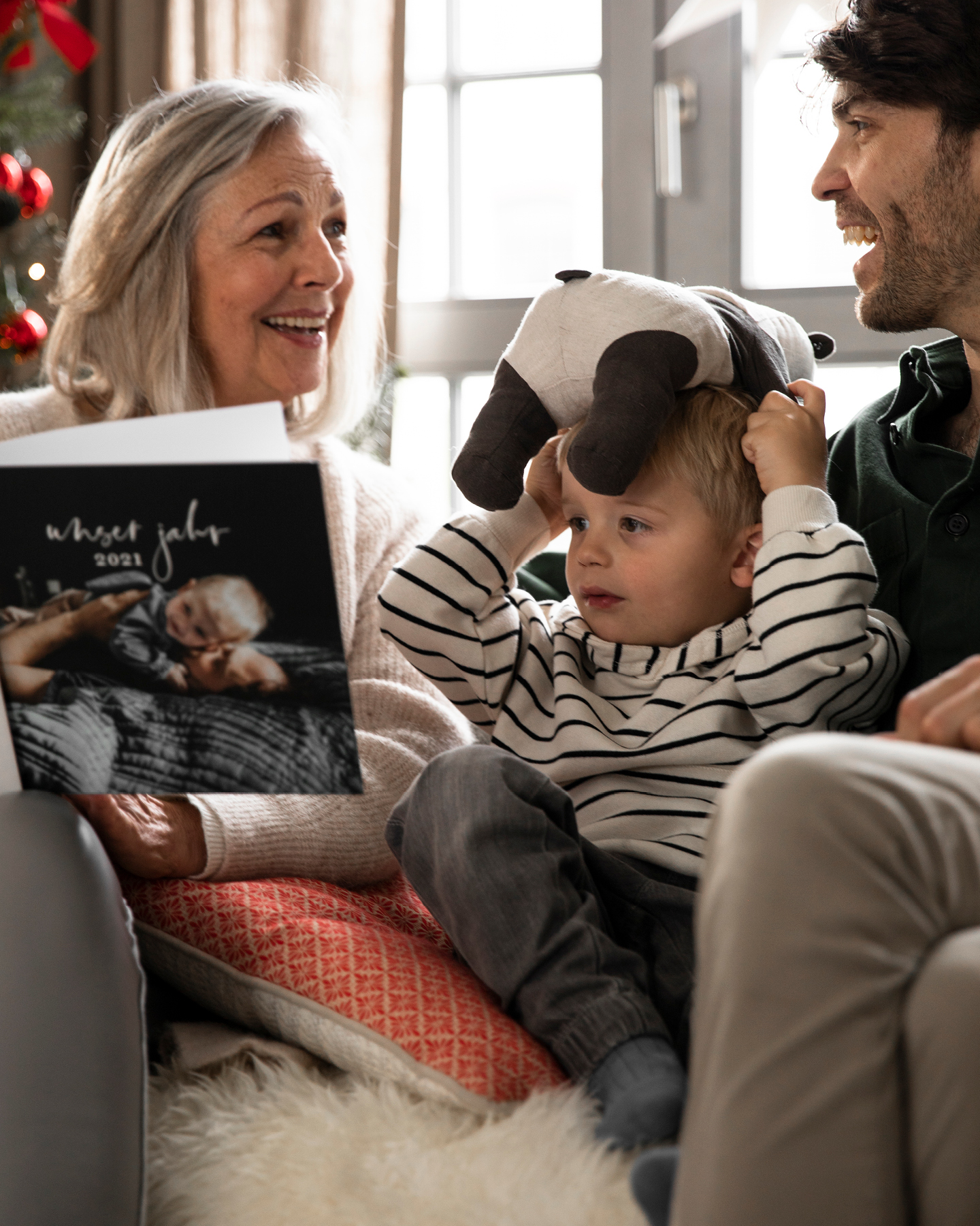 Eine Oma sitzt mit ihrem Enkel und dessen Vater auf der Couch und freut sich über ein Fotobuch zu Weihnachten