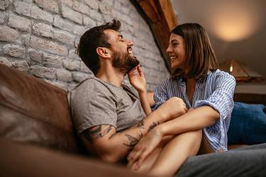  Zuhause auf dem Sofa schwelgen ein Mann und eine Frau in Vorfreude auf einen gemeinsamen Urlaub und schauen sich dabei lächelnd an.  