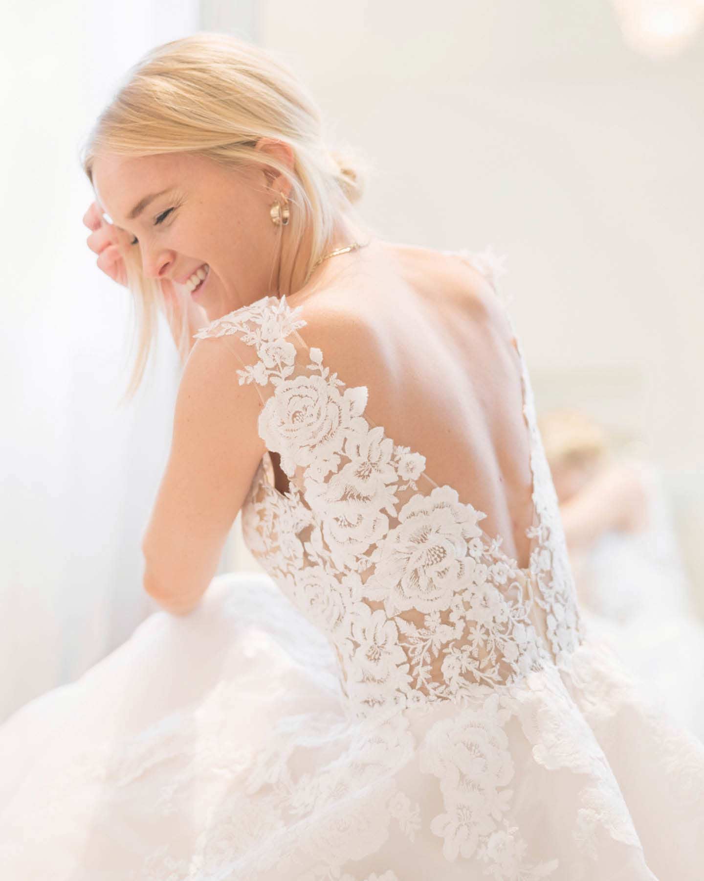 Braut lacht glücklich und zeigt den besonderen Rückenausschnitt mit Spitze ihres Brautkleides.