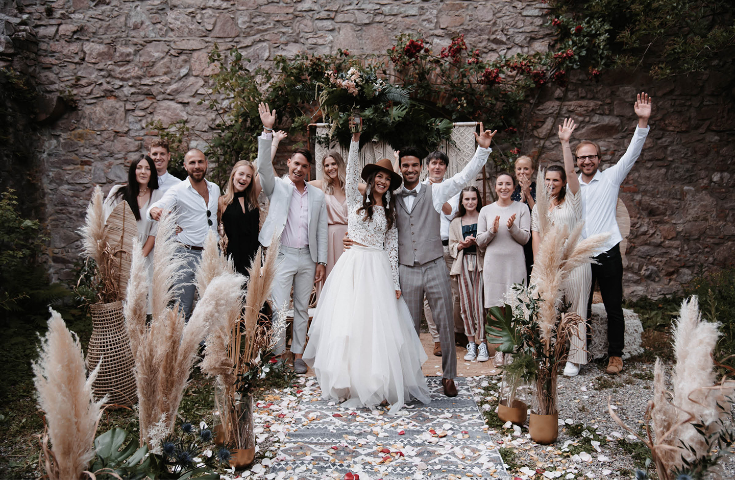 Hochzeitsbild mit Hochzeitspaar und Hochzeitsgesellschaft vor geschmückter Hochzeitslocation im Boho Stil.