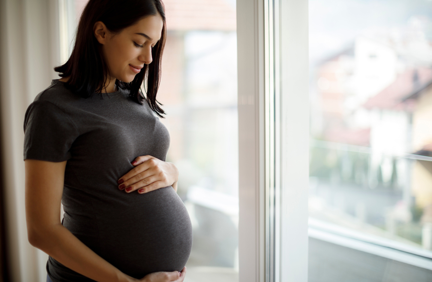 Hochschwangere vor Fenster umfasst liebevoll ihren Babybauch