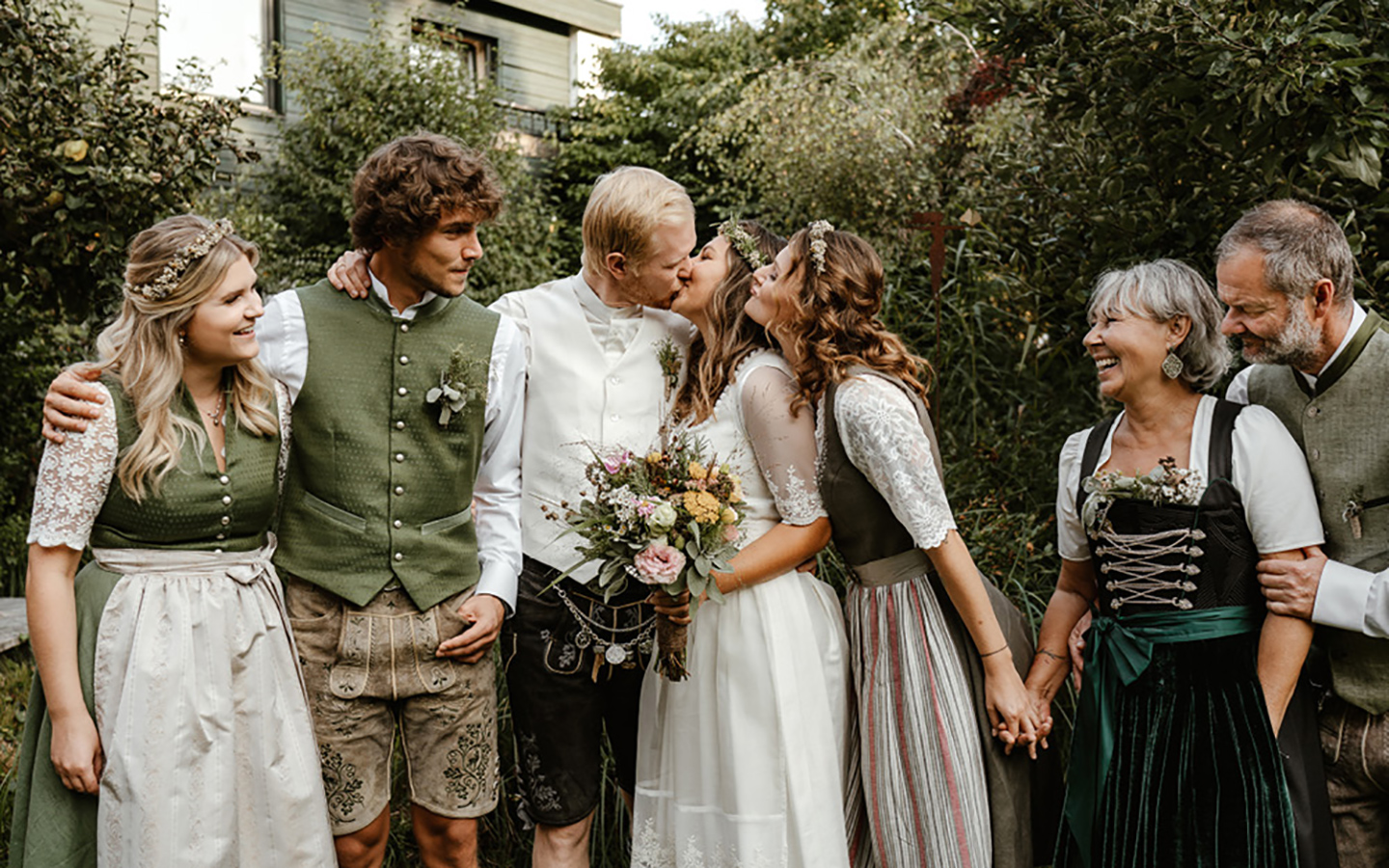 Die Gäste stehen zusammen im Garten, das Hochzeitspaar sich küssend in der Mitte. Alle anderen blicken zu ihnen und strahlen vor Freude über die Hochzeit. 