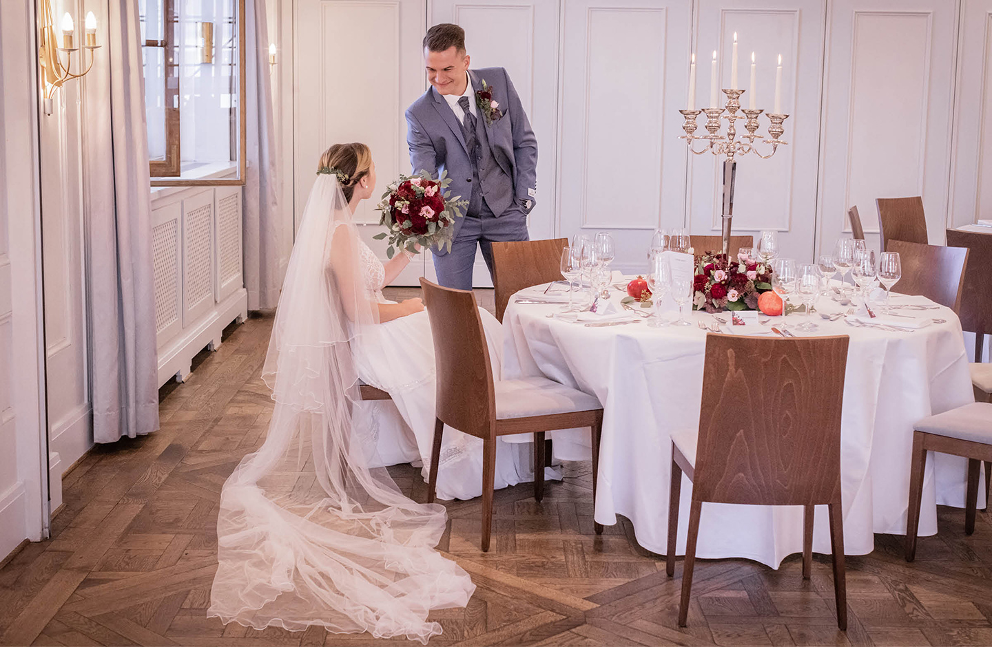 Braut und Bräutigam sehen sich zum ersten Mal in ihren Hochzeitsoutfits. Der Bräutigam trägt einen grau-blauen Anzug, die Braut ein weißes, schlichtes Kleid.