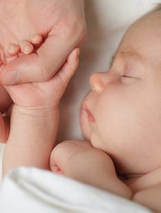 Nahaufnahme von Baby auf weißer Decke, seine Hand hält den Daumen des Vaters (nicht imBild)